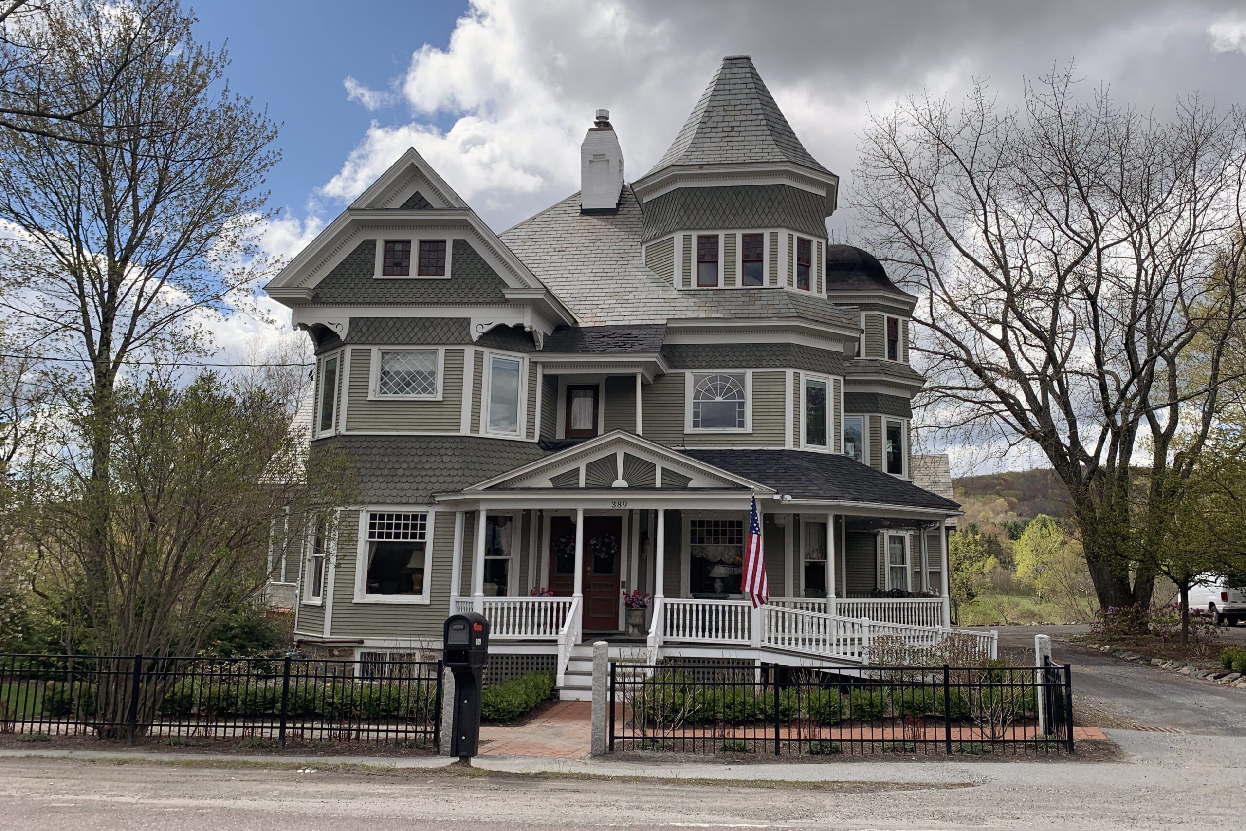 A large, three-story Victorian house with olive-green siding, white trim, and a prominent conical turret under a cloudy sky.