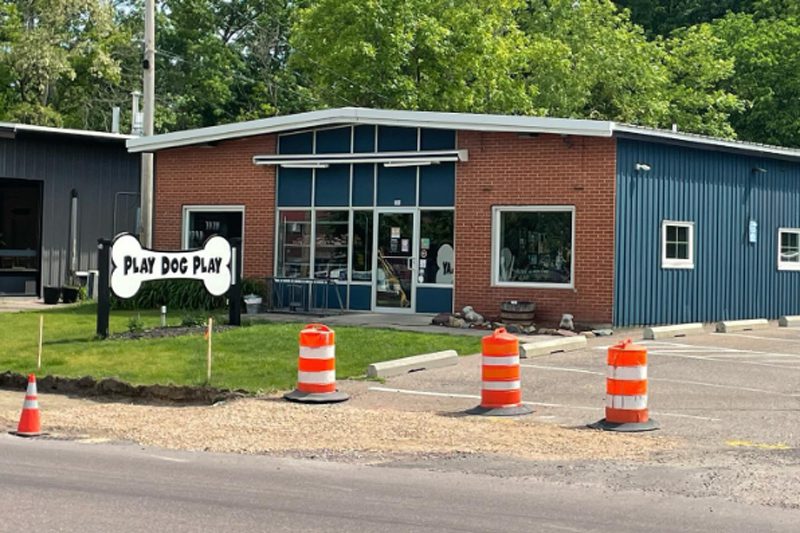 A single-story commercial building with a red brick facade and blue corrugated metal siding. A large white sign in the shape of a dog bone reads "PLAY DOG PLAY" on the front lawn. Three orange traffic barrels stand in the foreground.