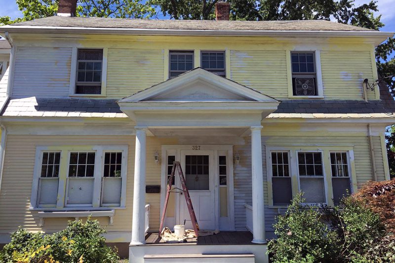 A yellow two-story house with white trim partially covered in patches of new white primer.